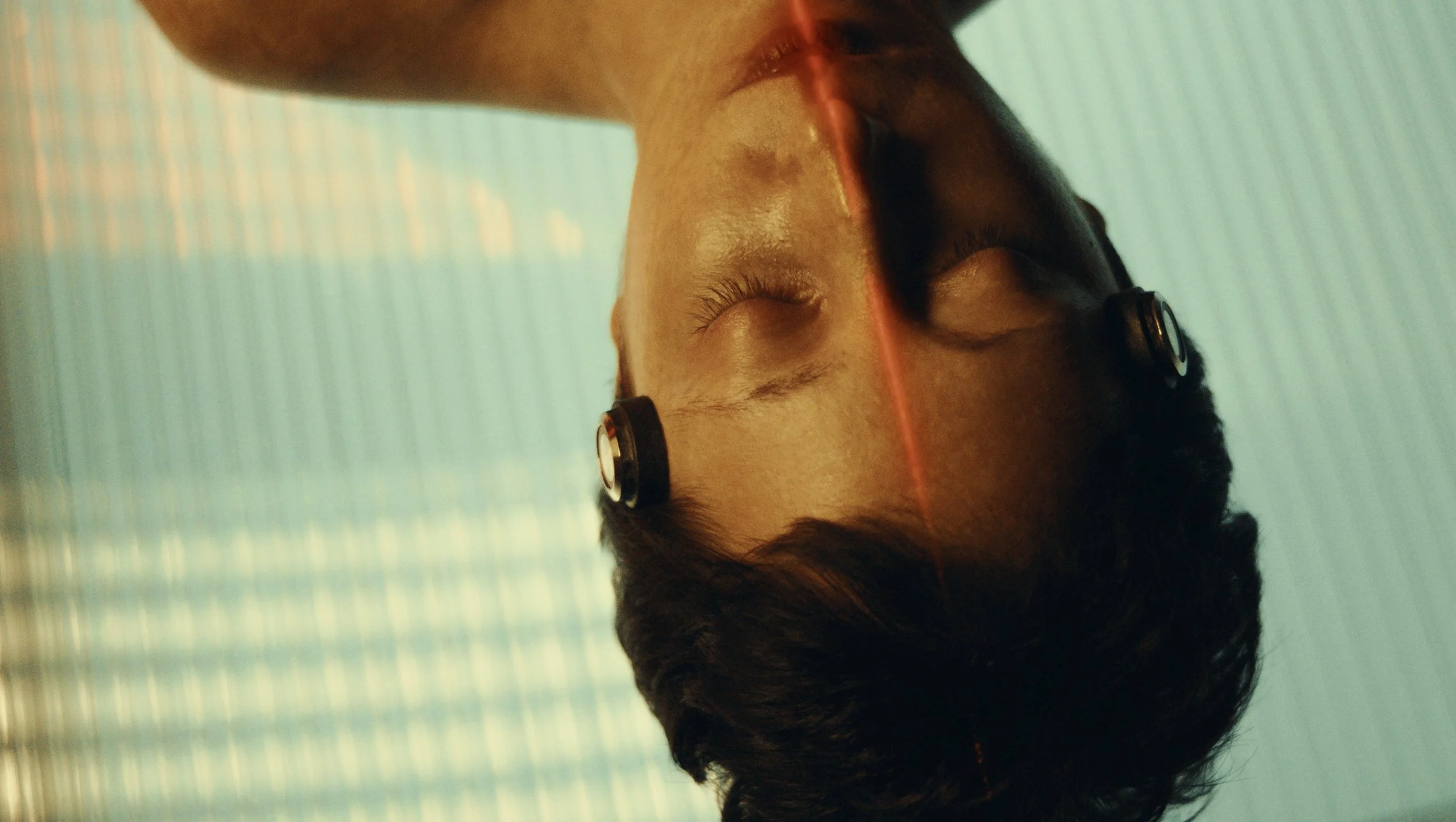 A young man with closed eyes, wearing wireless earbuds, lying in a swimming pool with water reflected on his face and a textured pool floor in the background.
