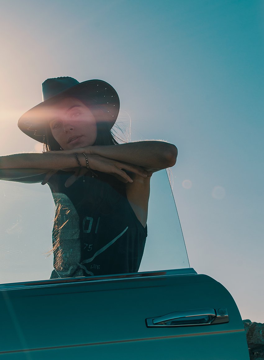 A woman wearing a wide-brimmed hat and dark tank top leaning on a car door with her arms crossed, backlit by the sun.