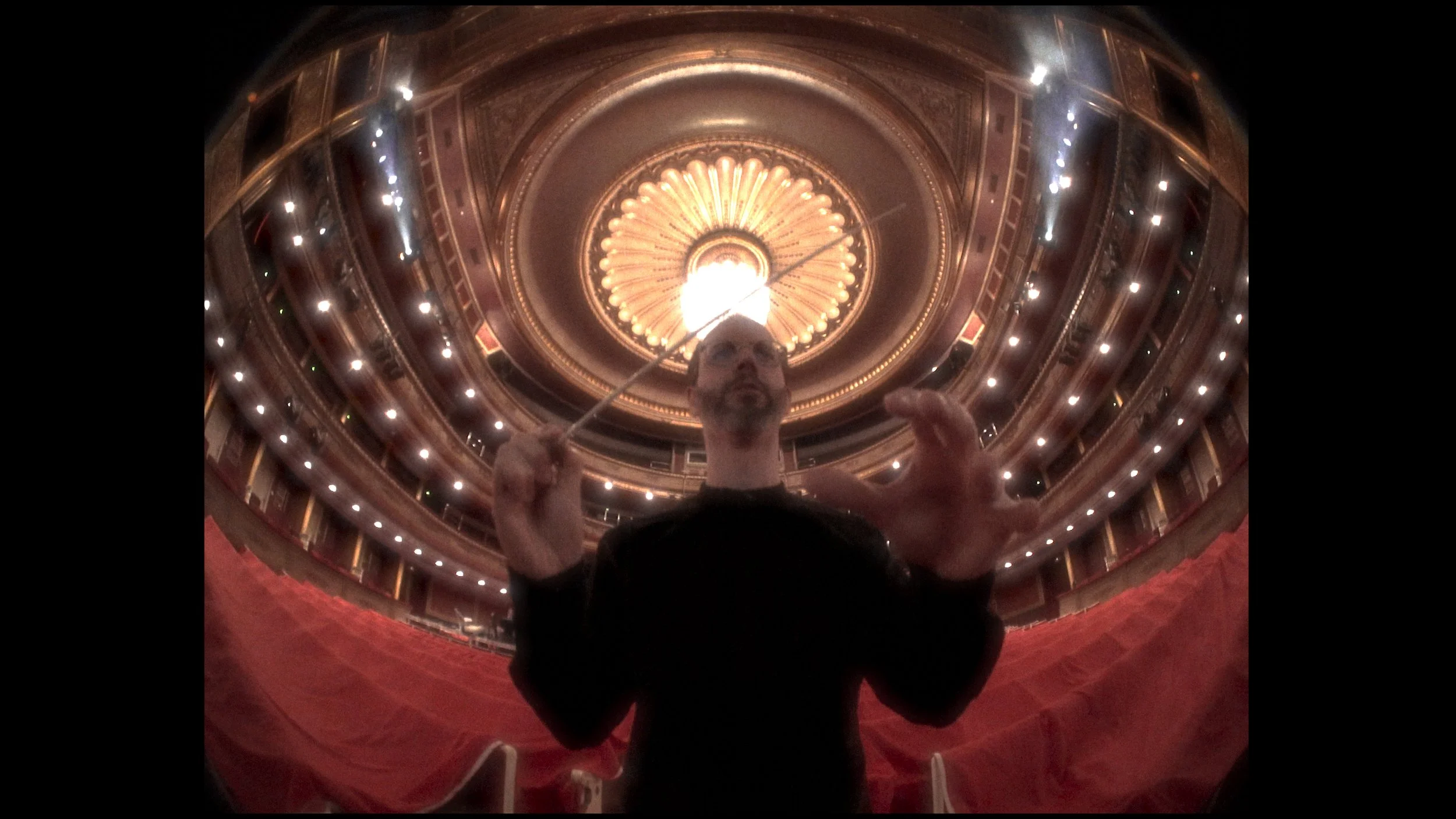 A man holding a conductor's baton standing on a stage inside a grand theater with a domed ceiling and ornate gold decorations, viewed from below.