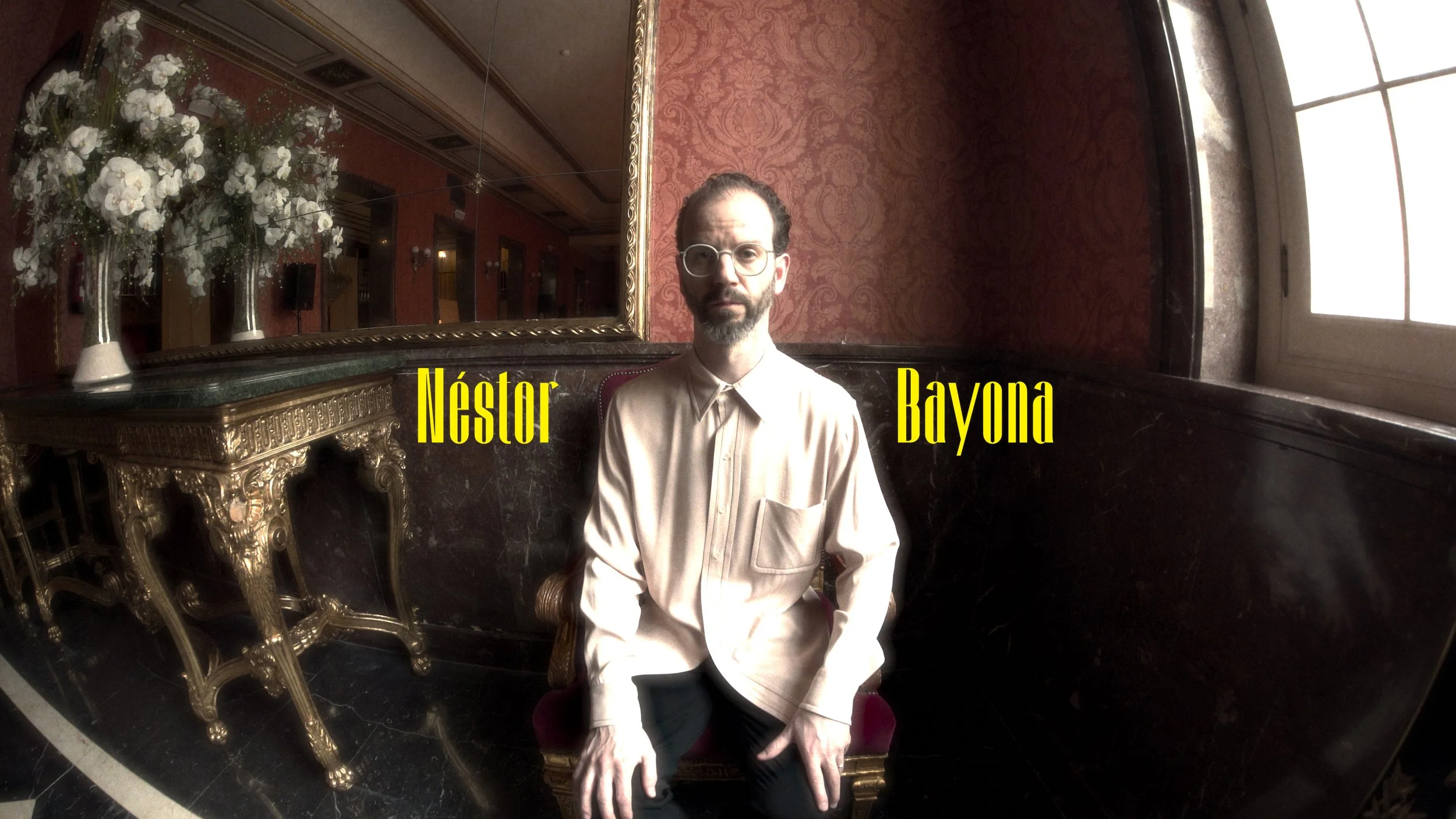 A man with glasses and a beard sits on a red velvet chair in an ornate room with dark marble walls, gold decor, and a mirror. Two vases with white flowers sit on a gilded console table behind him. The man wears a light-colored shirt and black pants.