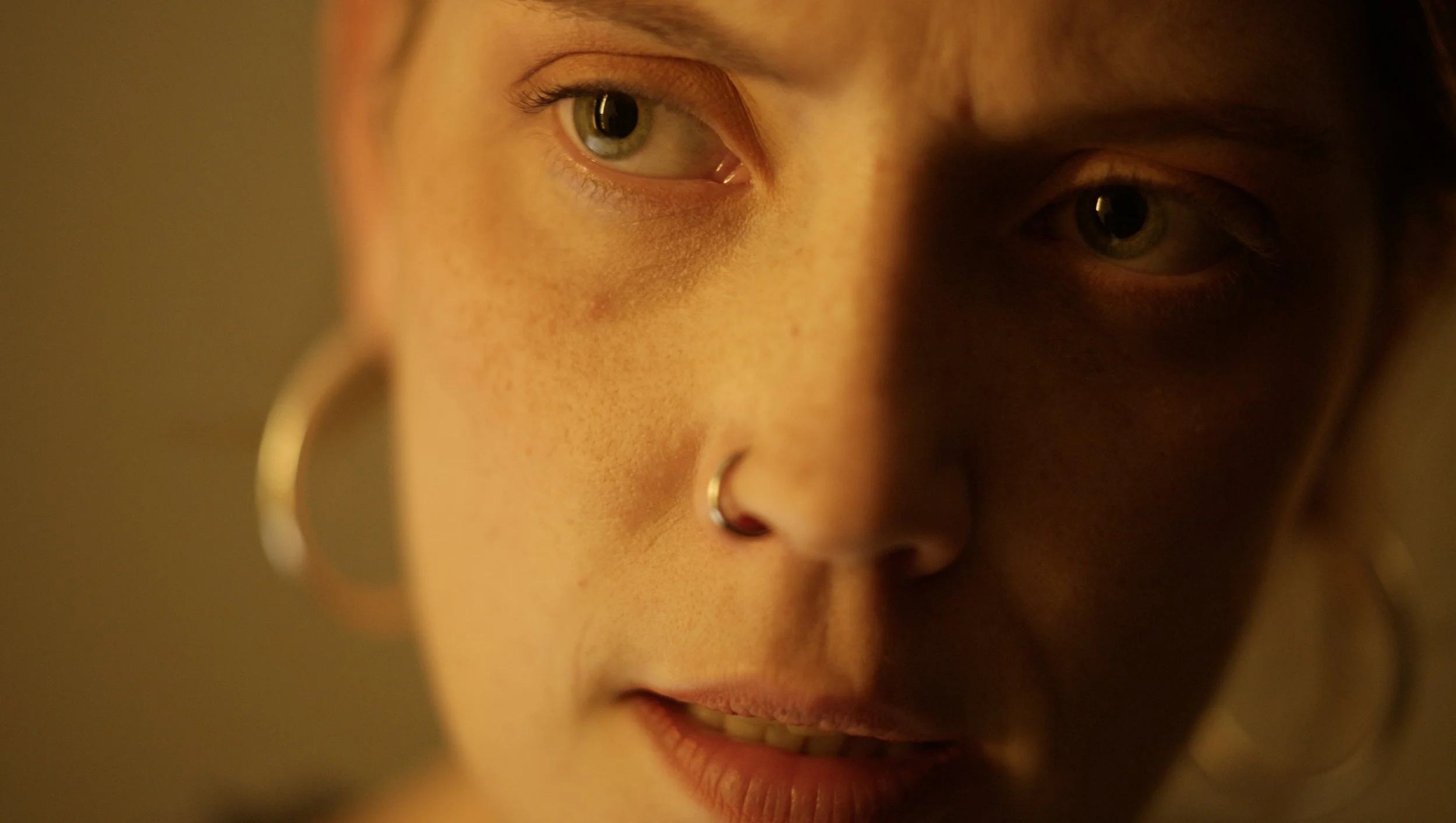 Close-up of a young woman's face showing one eye, part of her nose with a hoop nose ring, and lips, with warm lighting and soft focus.