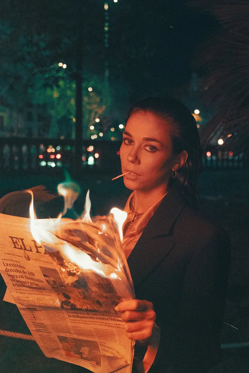 A woman in a dark suit smoking a cigarette while holding a burning newspaper outdoors at night.