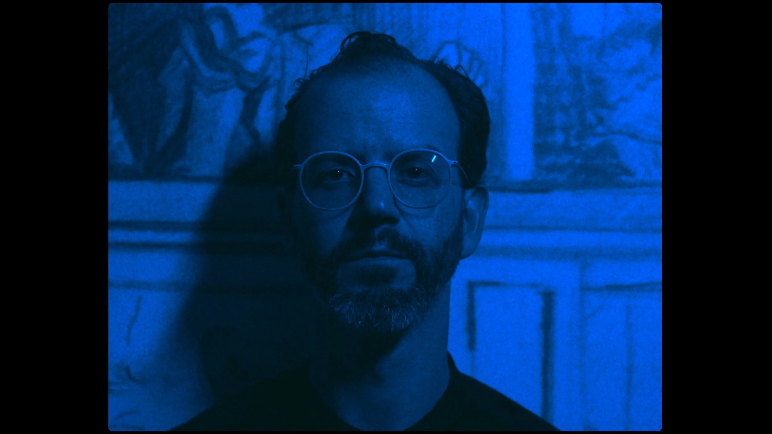 A man with glasses and facial hair lies down on a surface, illuminated by blue lighting, with artwork visible in the background.