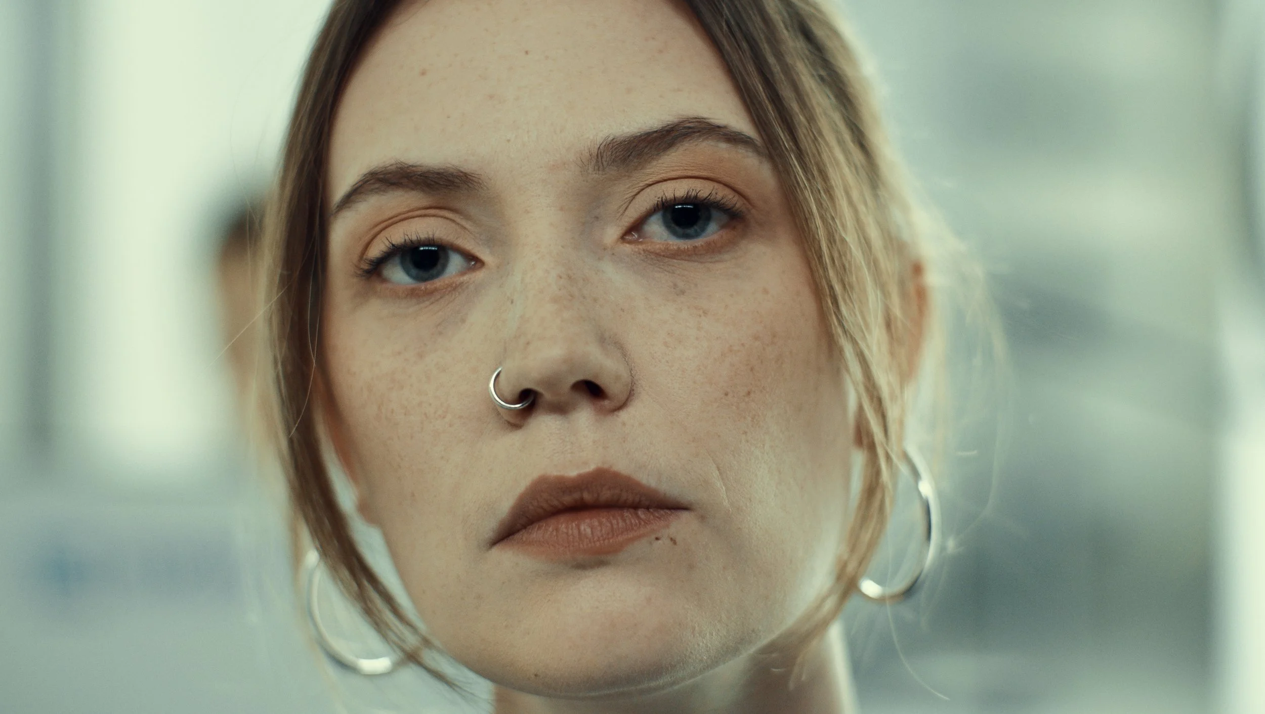Close-up of a young woman with blue eyes, freckles, a nose ring, and hoop earrings, looking directly at the camera with a neutral expression.