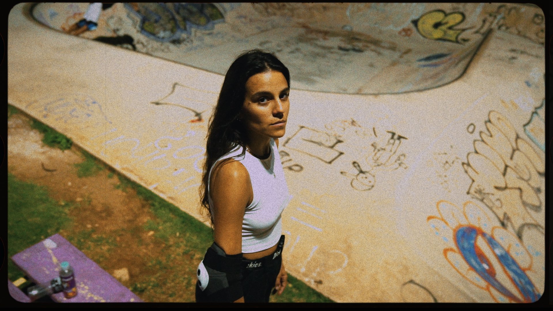 A young woman with long dark hair looks up at the camera while standing outdoors in front of a concrete wall covered with colorful graffiti and drawings.