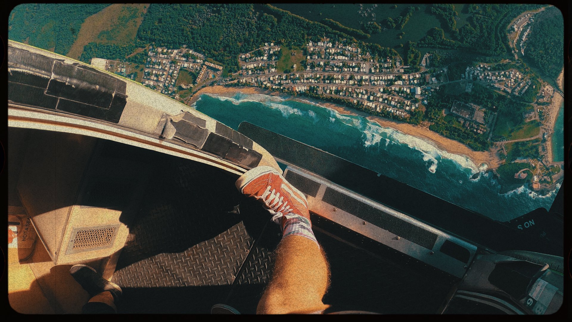 Inside a helicopter cockpit showing a person's legs, with a view of a coastal city and beach below through the window.