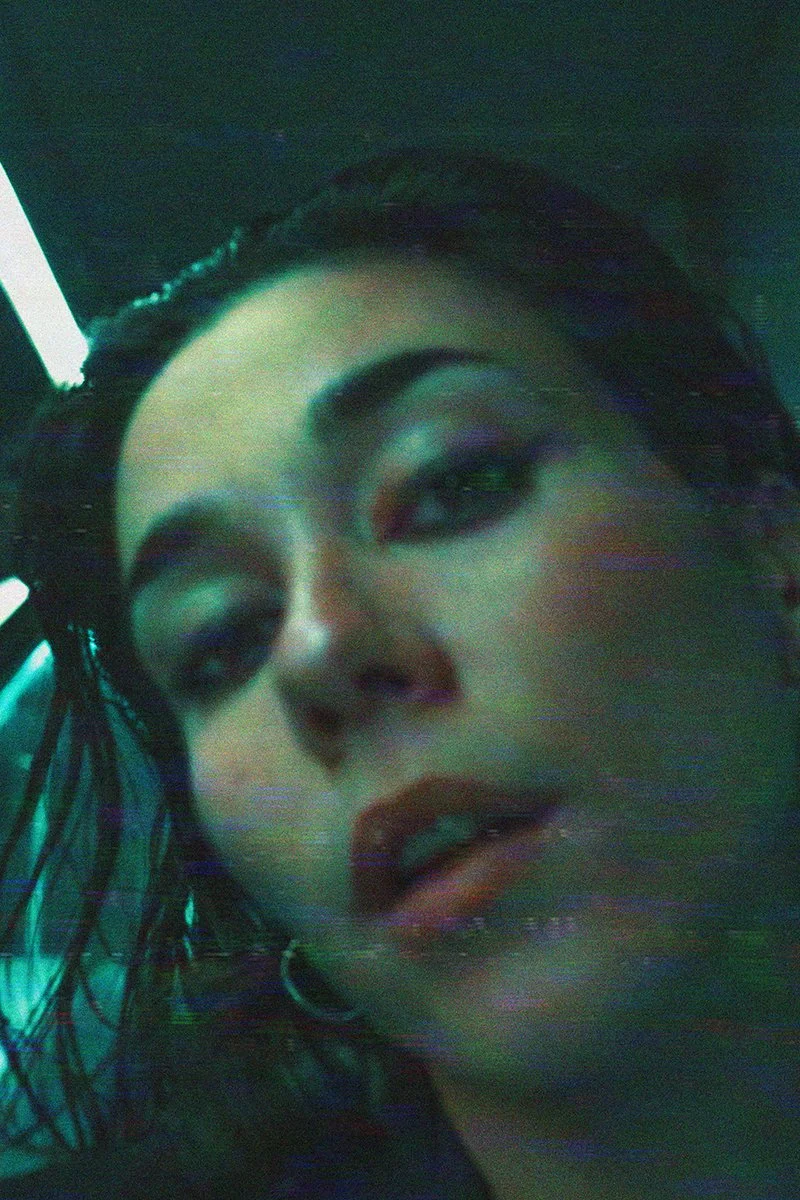 Close-up of a woman's face with makeup and wet hair, with a dark background and fluorescent lighting.