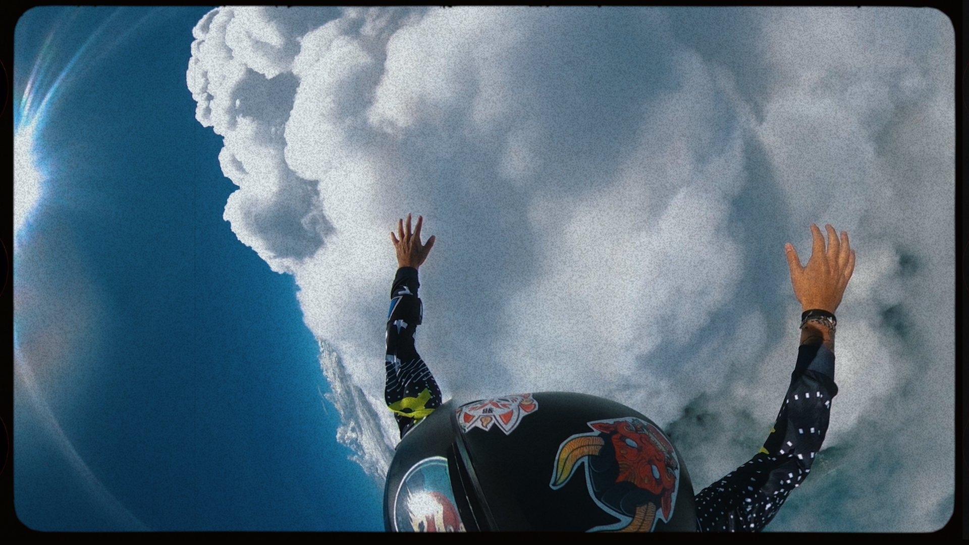 A person wearing a black helmet with colorful dragon artwork and a black and white patterned shirt, arms raised, appears to be skydiving or free-falling through a sky filled with large clouds and blue sky.