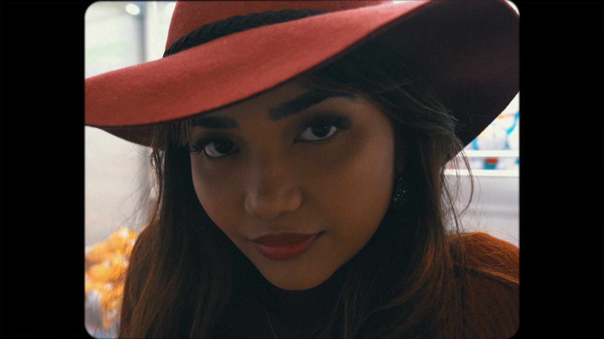 A woman with long dark hair wearing a large brown hat with a black band, looking directly at the camera with a slight smile, wearing red lipstick and earrings.