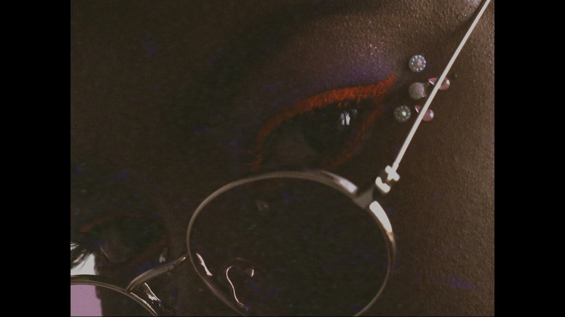 Close-up of a makeup eye with red eyeshadow, black eyeliner, and small decorative rhinestones on the eyelid, photographed in low light with reflections and a mirror.
