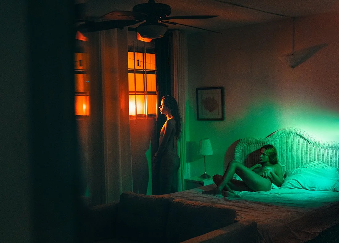 Two women in a dimly lit bedroom; one standing near the window, the other lying on the bed.
