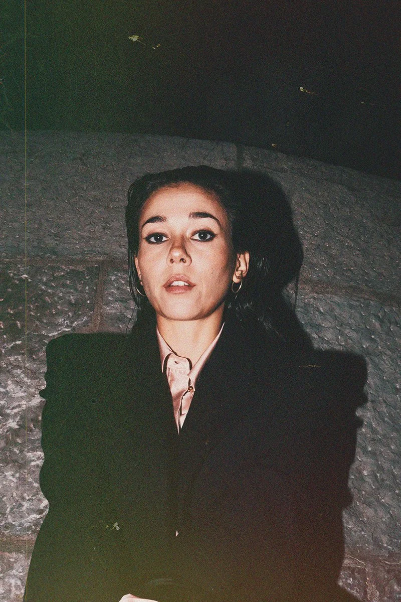 A young woman with dark hair, wearing a black blazer and pink shirt, sits against a stone wall in a dimly lit environment, looking directly at the camera.