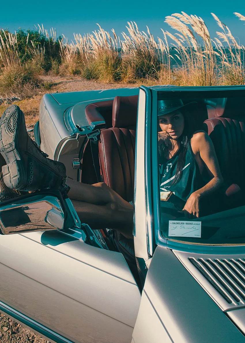 A woman with a cowboy hat sitting inside a white convertible car with maroon interior, parked outdoors near tall grass and a clear blue sky.