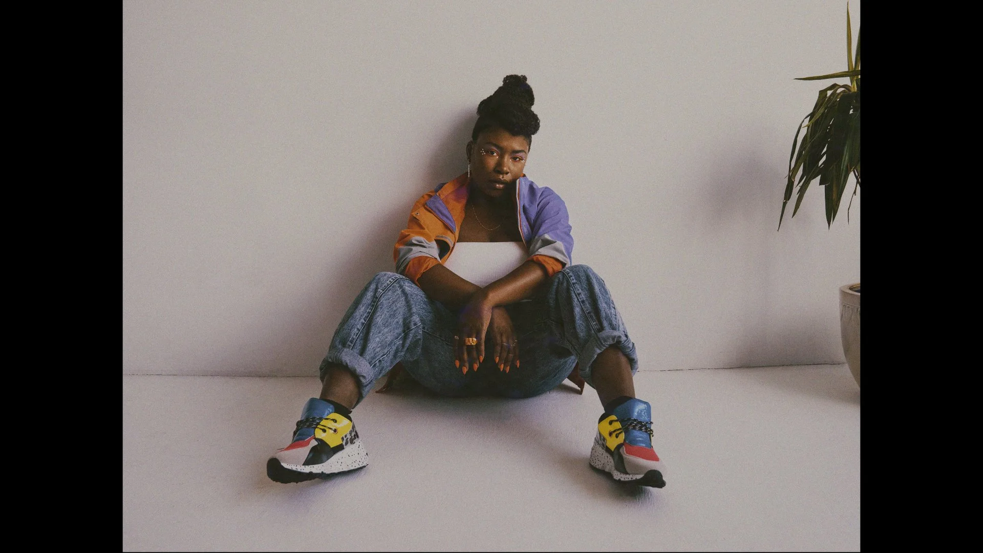 Young woman sitting on the floor against a light gray wall, wearing oversized jeans, a colorful windbreaker, and multicolored sneakers, with a potted plant visible on the right side.
