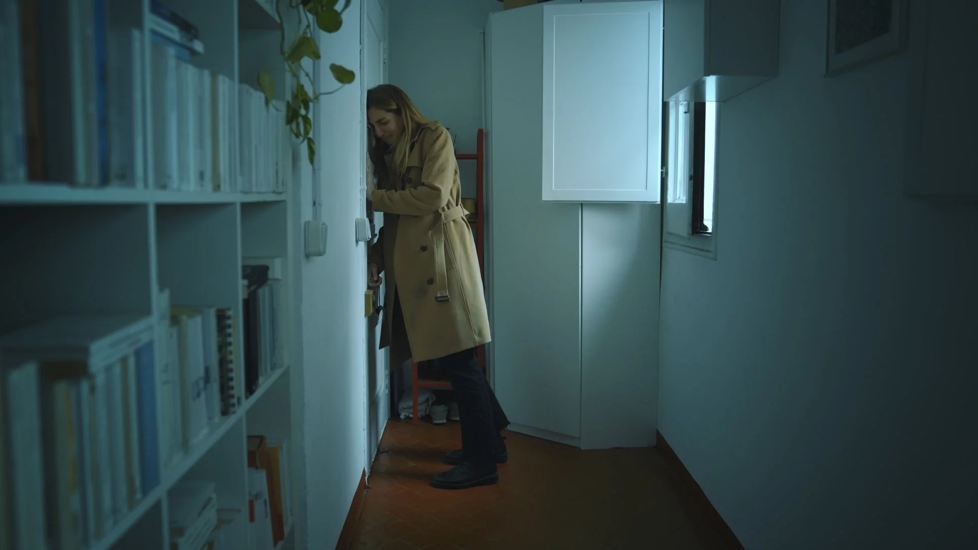 A woman wearing a beige trench coat and black pants stands in a narrow room, reaching towards a doorbell or intercom on the wall, with a slight smile. The room has white walls, a wooden floor, a tall white cabinet, a small window, and a bookshelf fil