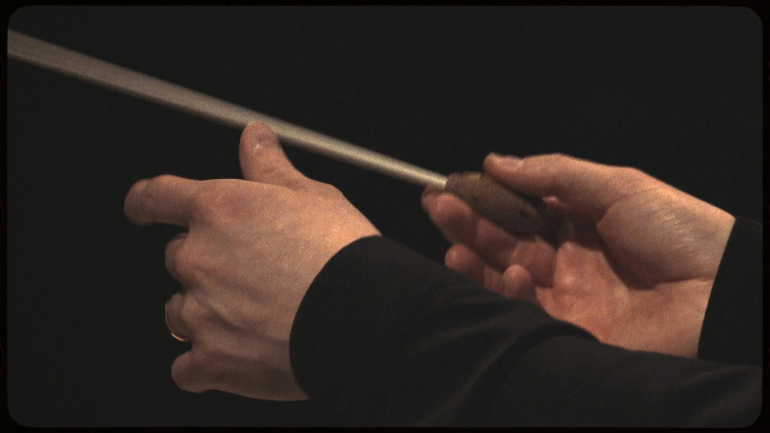 Close-up of hands holding a small tool and a thin rod, with a dark background.