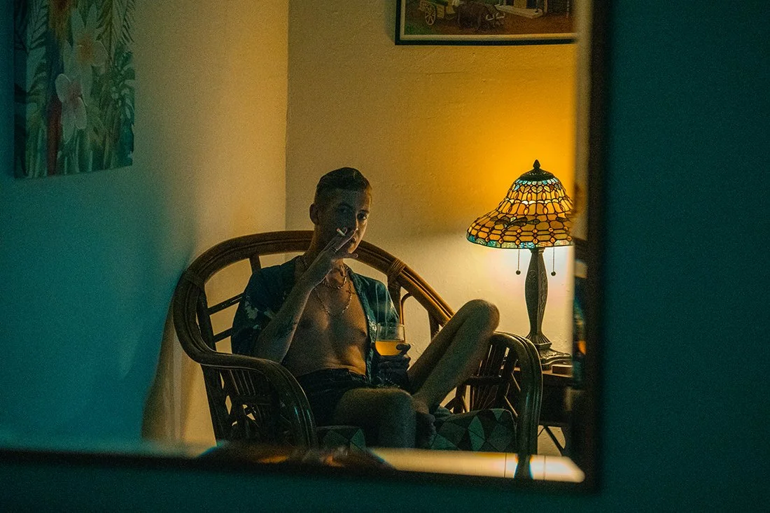 A young man sitting in a wicker chair in a dimly lit room, smoking a cigarette and holding a glass of orange drink. There is a table lamp with a stained glass shade emitting a warm glow, and artwork on the wall.