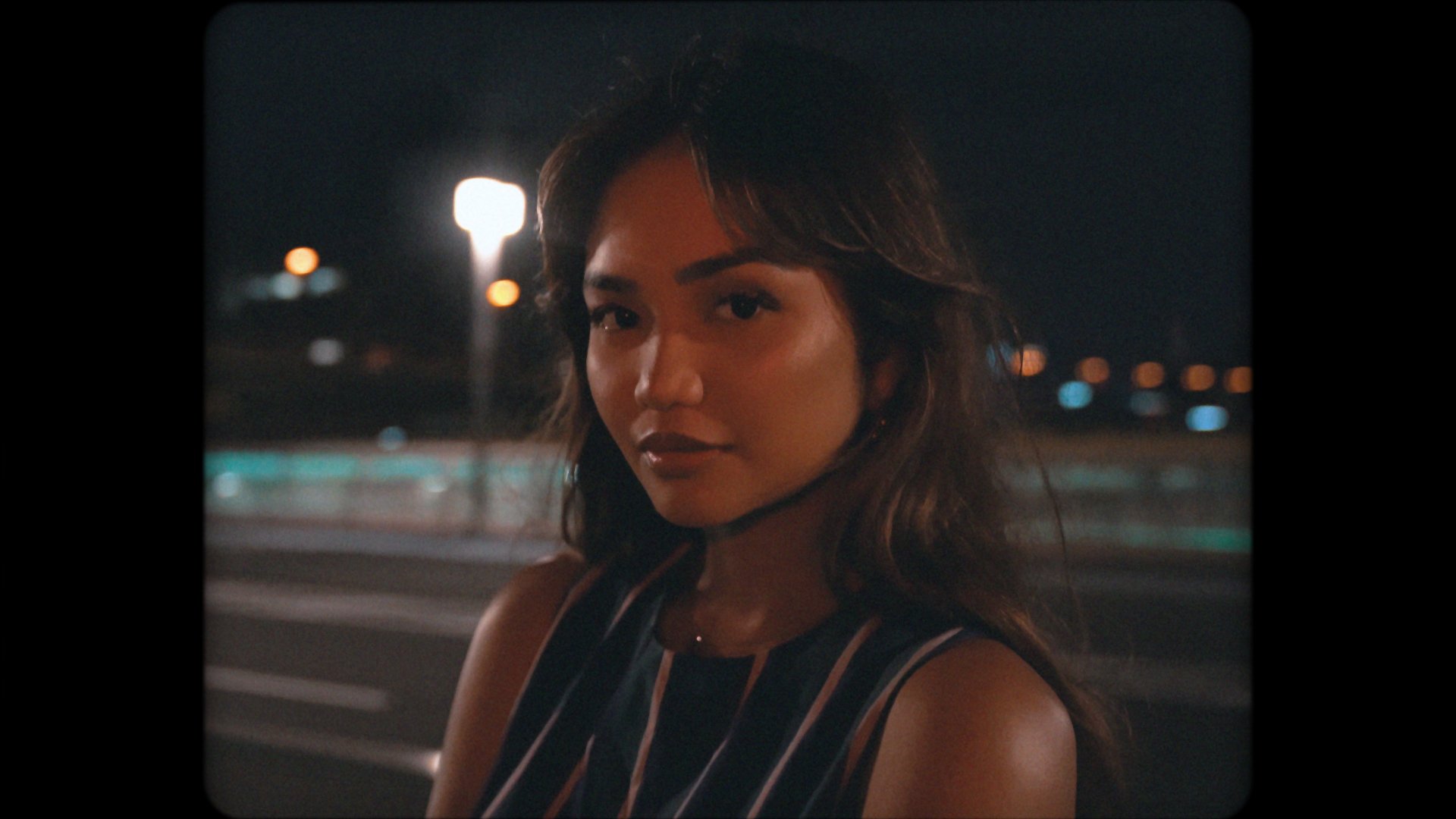A woman with wavy brown hair and wearing a sleeveless striped top poses at night with city lights in the background.