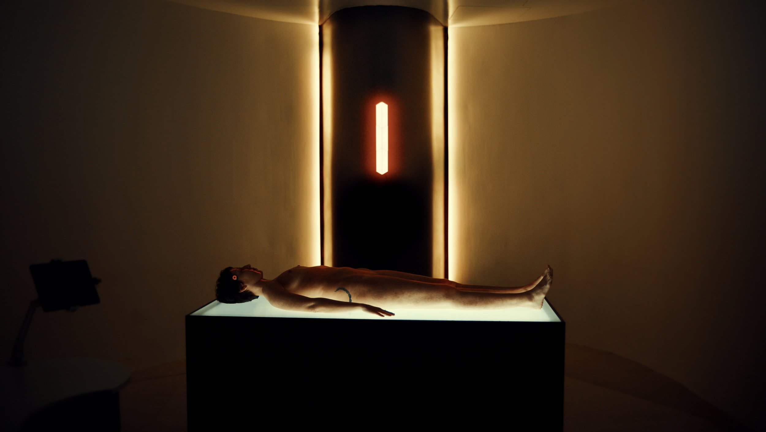Nude mannequin lying on back on illuminated display table in a dark room, with a black door and red light in the background.
