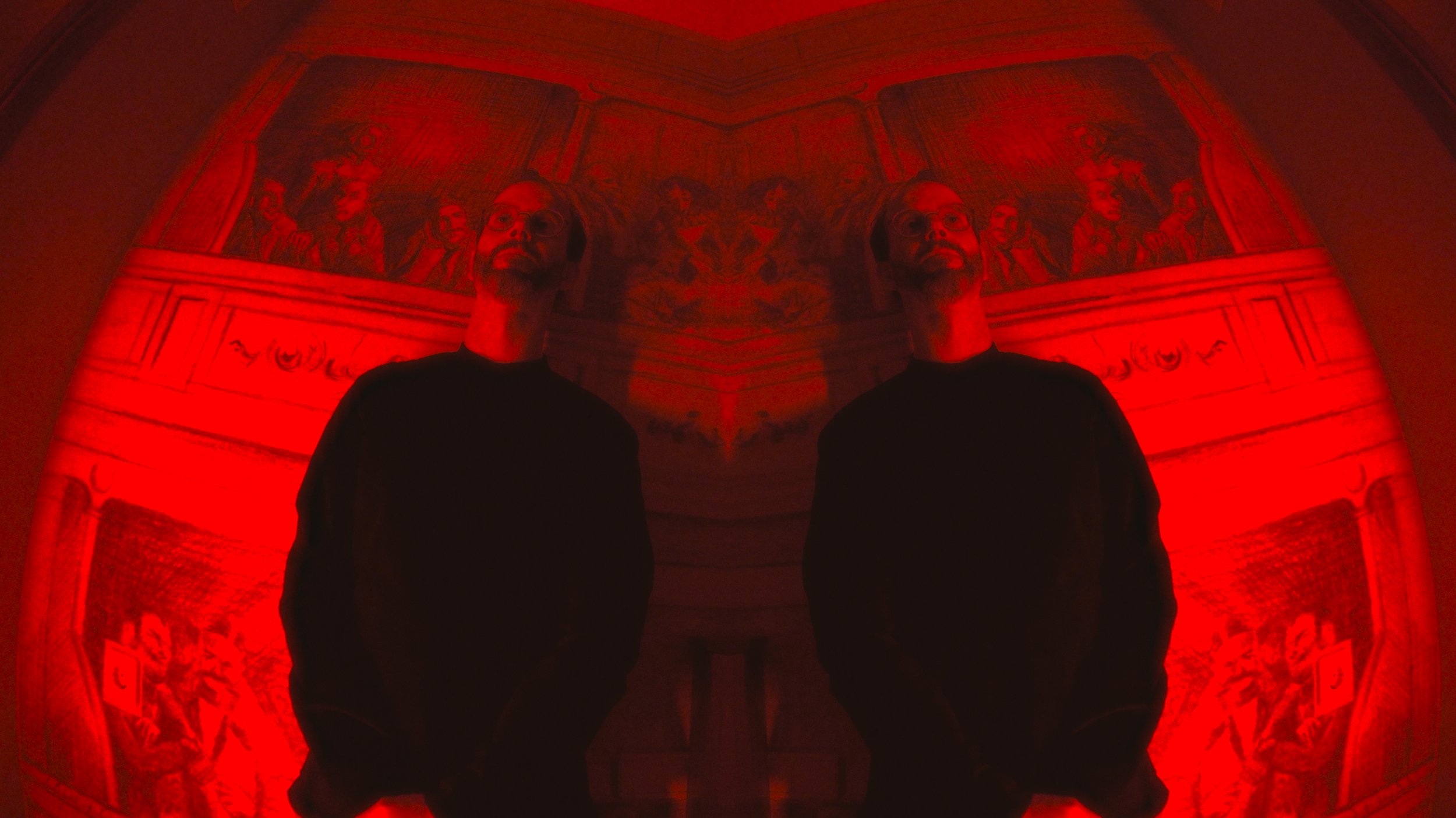 A person with glasses and a beard in a dark room illuminated by red light, with a mirror creating a symmetrical reflection, revealing a painted ceiling with detailed artwork.