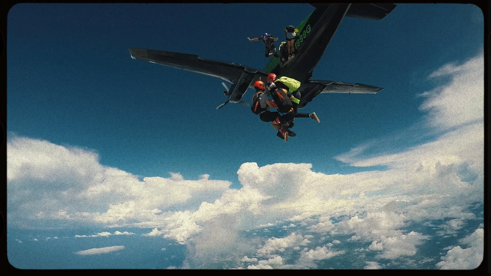 Skydivers exiting an airplane above the clouds with a clear blue sky.