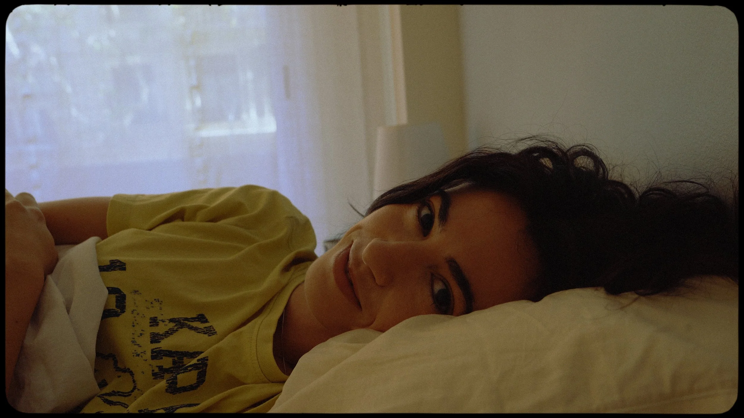 Woman lying on her side in bed, smiling and looking at the camera, with dark curly hair and wearing a yellow T-shirt.