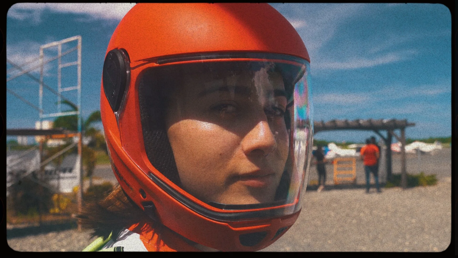 Woman wearing an orange motorcycle helmet with a clear visor, outdoors on a sunny day, with people and a wooden structure in the background.