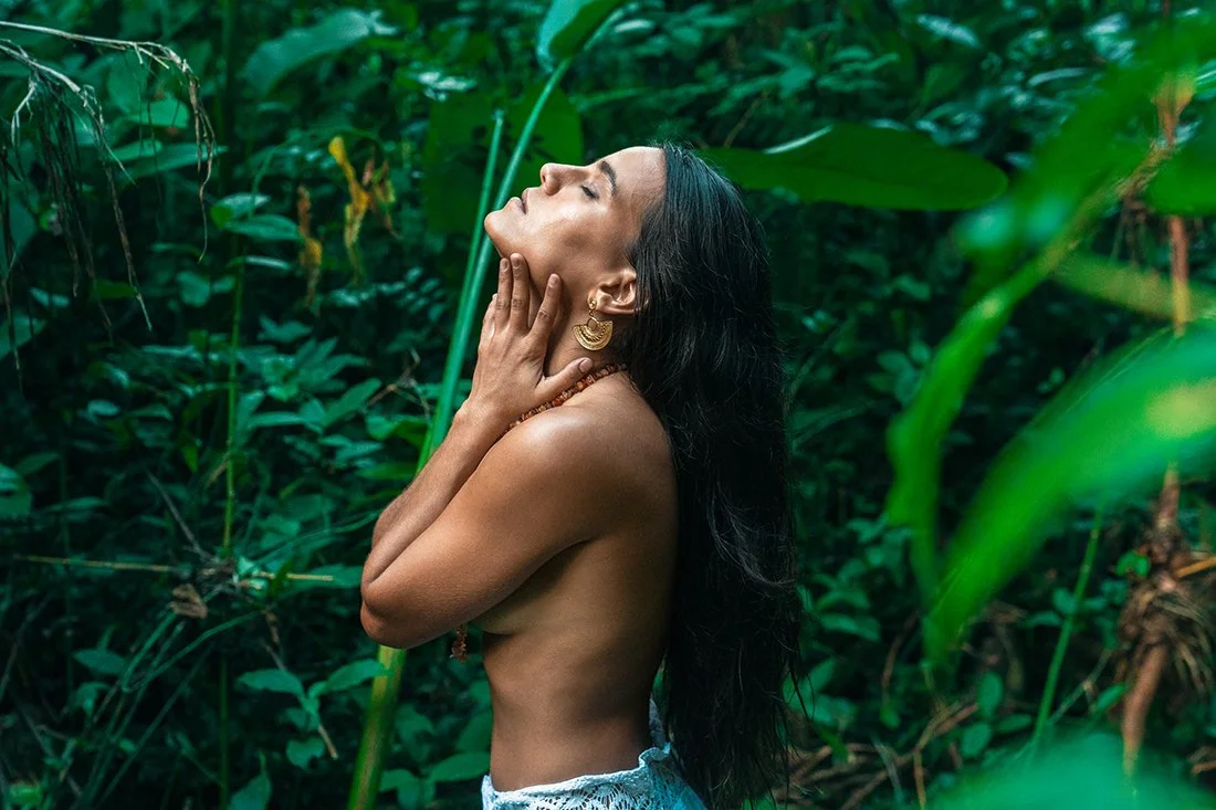 A woman with long black hair and gold jewelry standing topless in a lush green jungle, with her eyes closed and hand gently touching her neck.