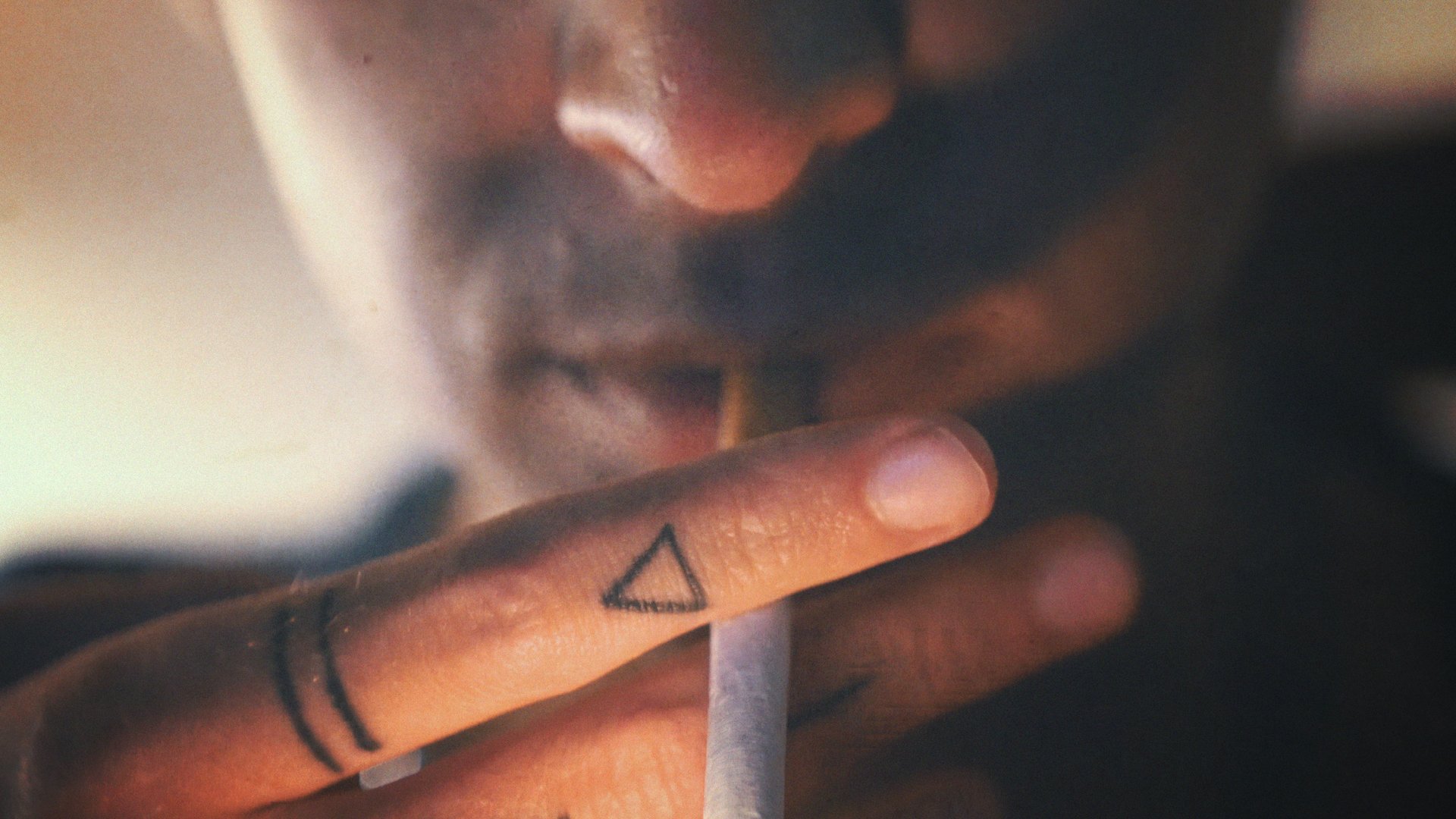 Close-up of a person's face with a cigarette near their lips, showing a tattoo of a triangle on their finger and a ring on their finger.