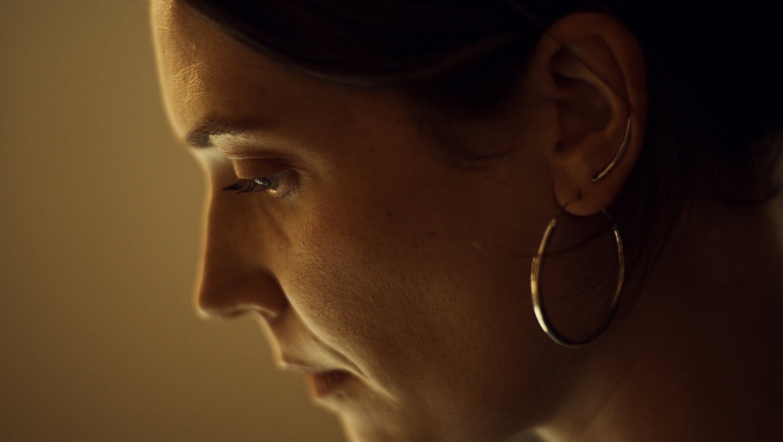 Close-up side profile of a woman's face in low lighting, wearing hoop earrings and looking downward.