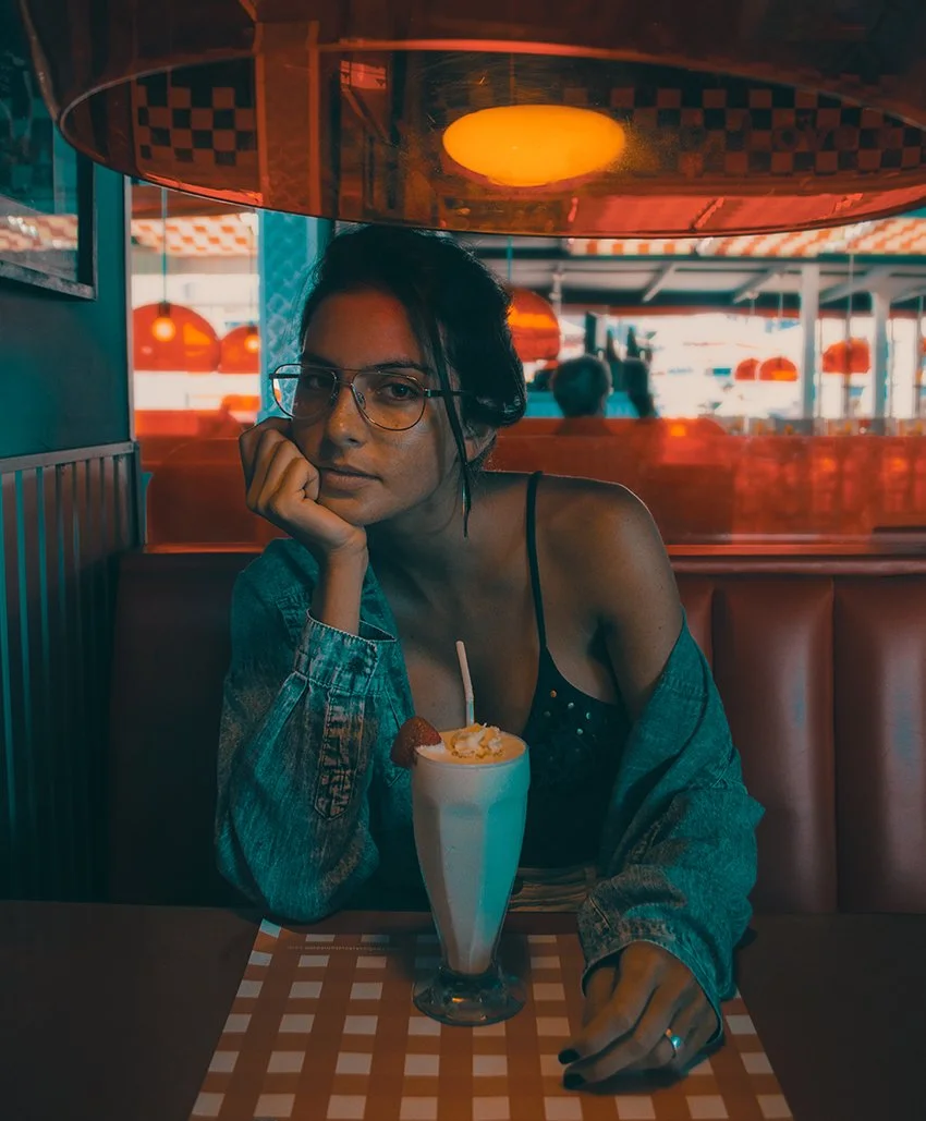 Young woman with glasses sitting at a diner booth with a milkshake topped with whipped cream and a strawberry, resting her chin on her hand and looking at the camera.