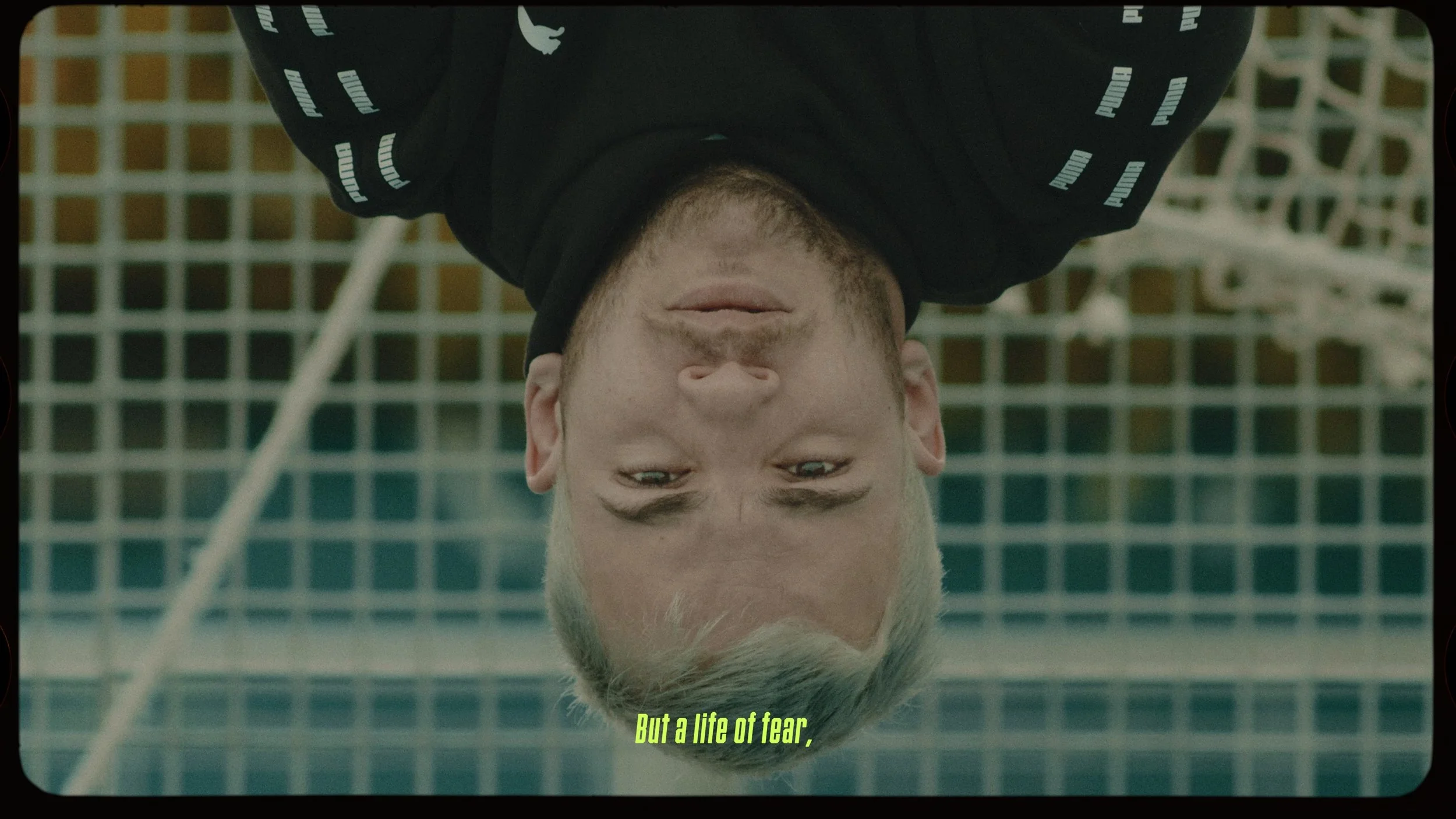 Upside-down image of a man with blond hair, looking directly at the camera, with a grid-like pattern and metal structure in the background. Yellow caption text reads, 'But a life of fear,'.