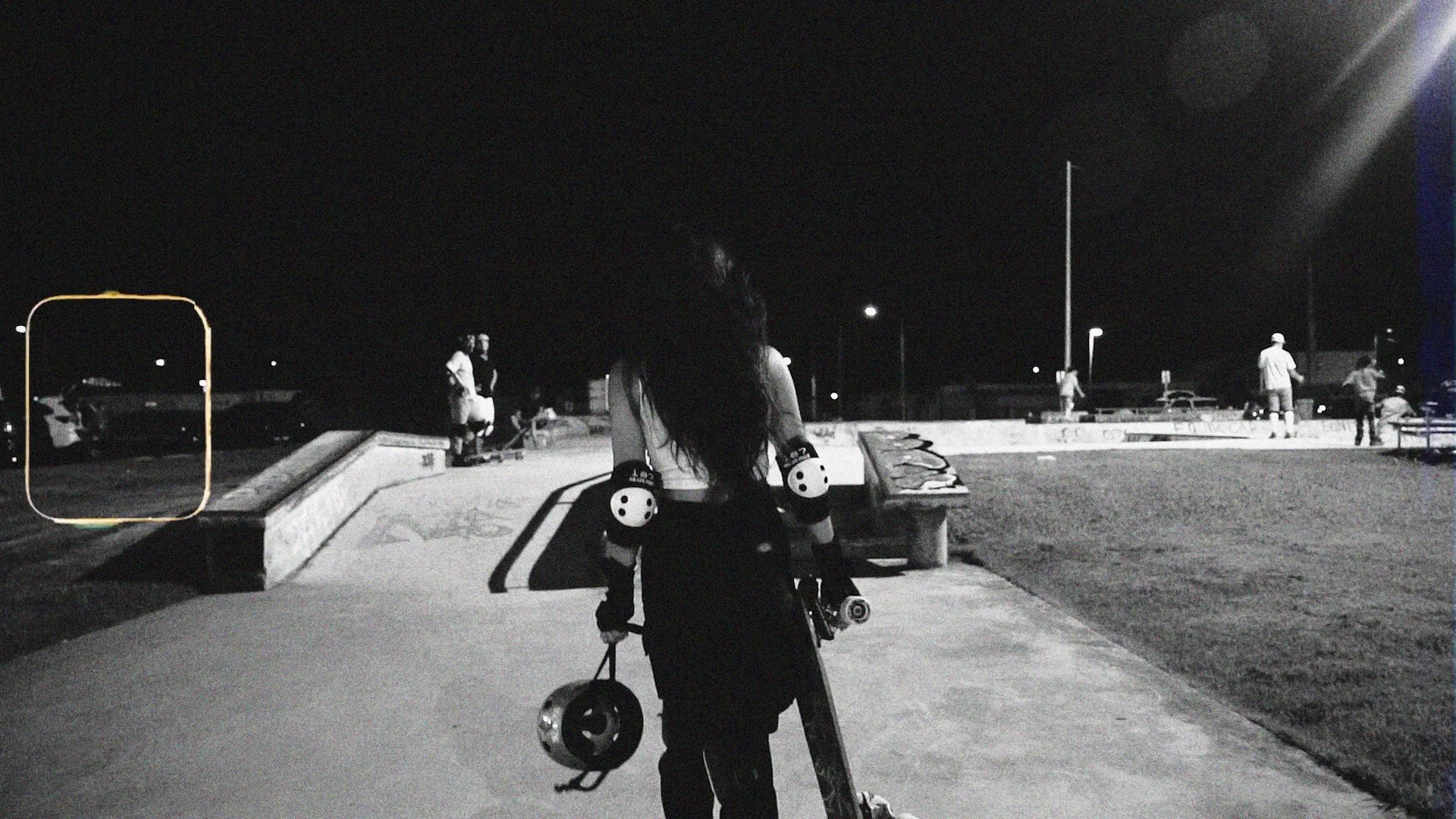 A woman walking at night in a skate park, holding a helmet and skateboard, wearing protective knee pads, with other skaters in the background.
