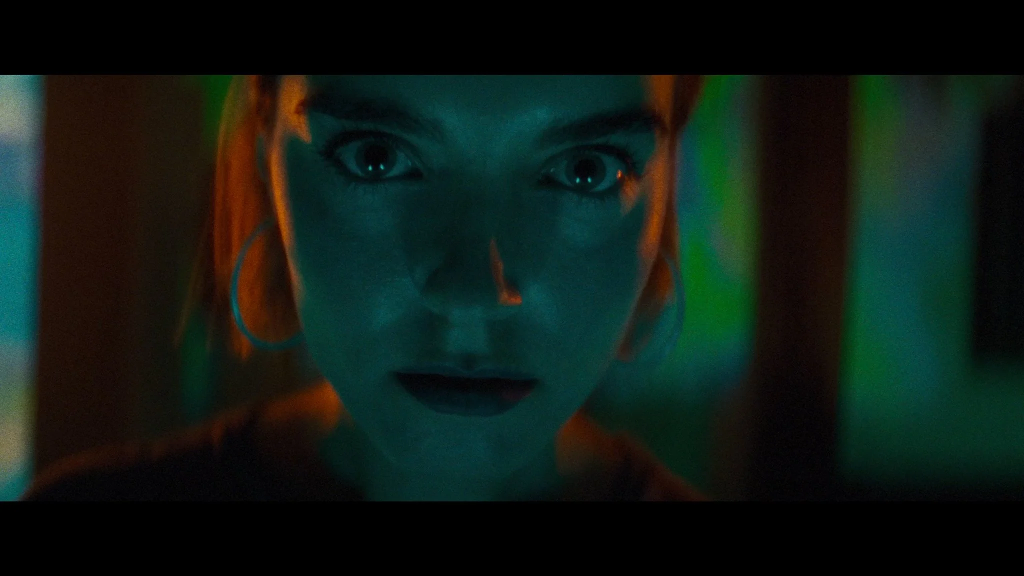 Close-up of a woman with wide eyes and dark lipstick, illuminated by green and orange light.