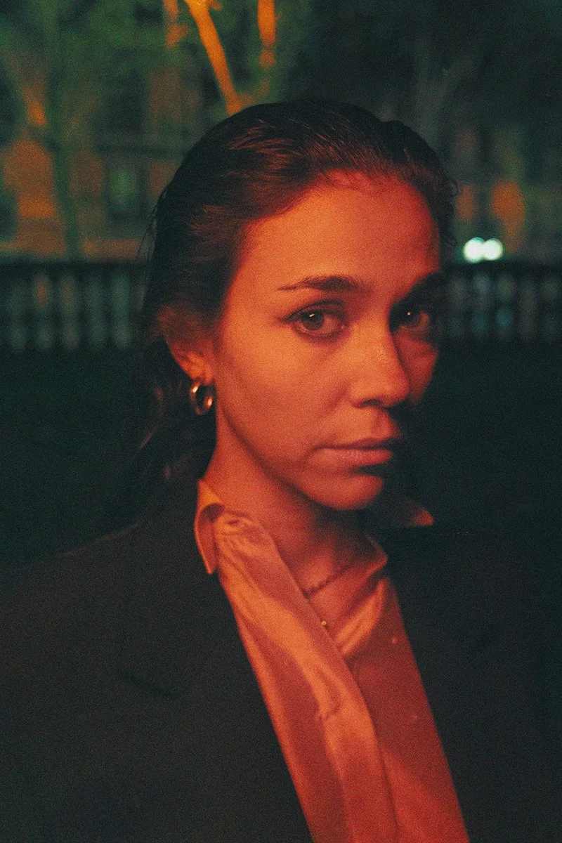 A woman with dark wet hair, wearing earrings and a black blazer, is in a dimly lit setting with orange and green lighting.