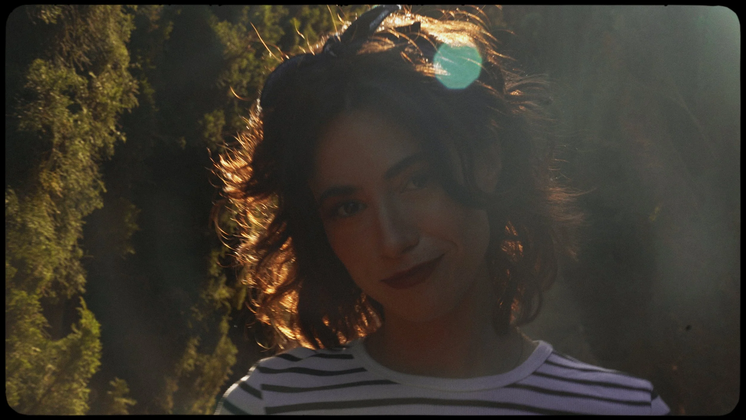 A woman with curly brown hair, wearing a striped shirt, smiling outdoors with sunlight and trees in the background.