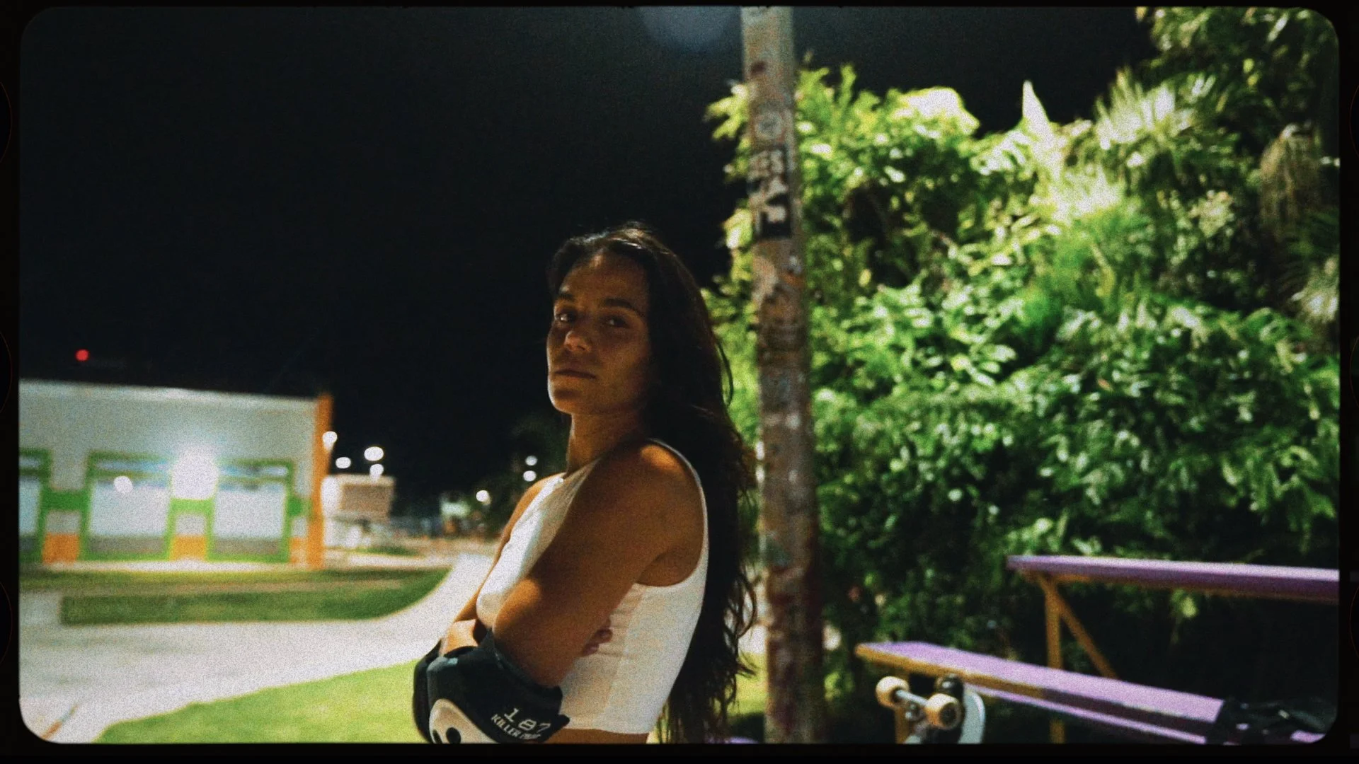 A woman standing outdoors at night with her arms crossed, wearing a sleeveless white top and black gloves, with a skateboard sitting on a bench to her right, and lush green foliage behind her.