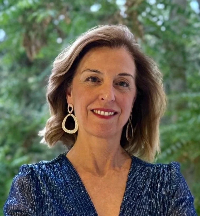 A woman with shoulder-length blonde hair wearing a blue sparkly dress and large drop earrings, smiling outdoors with green foliage in the background.