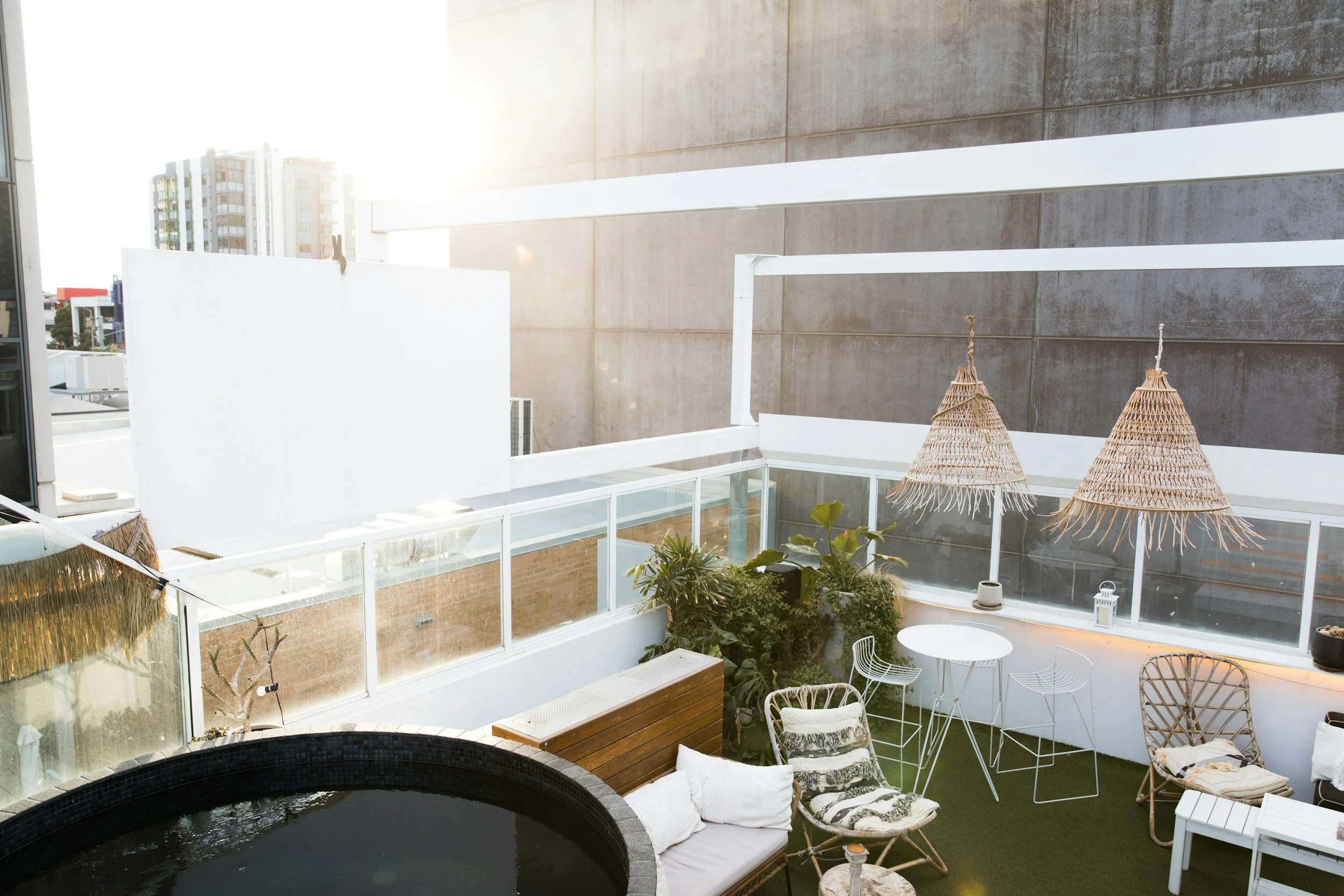 Sunlit balcony with wicker umbrellas, white chairs, and potted plants, including a black hot tub and outdoor furniture against city buildings.