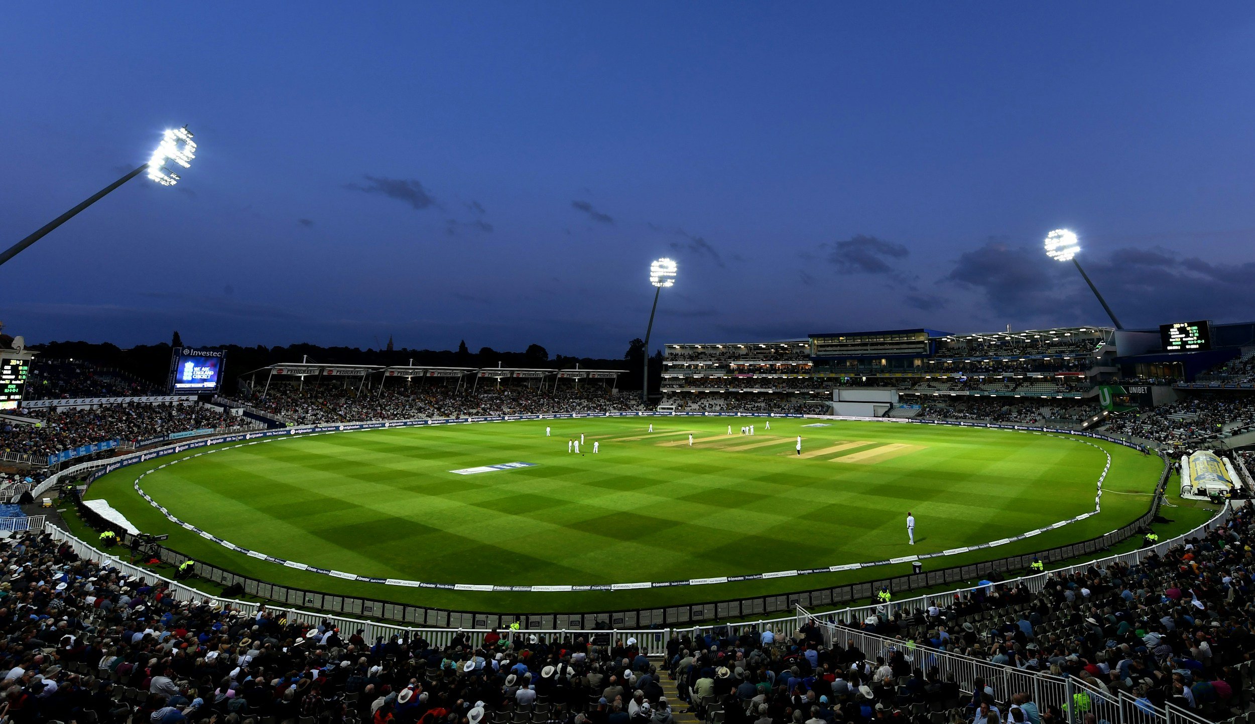 Cricket stadium during evening with players on the field, audience in stands, and floodlights illuminating the field