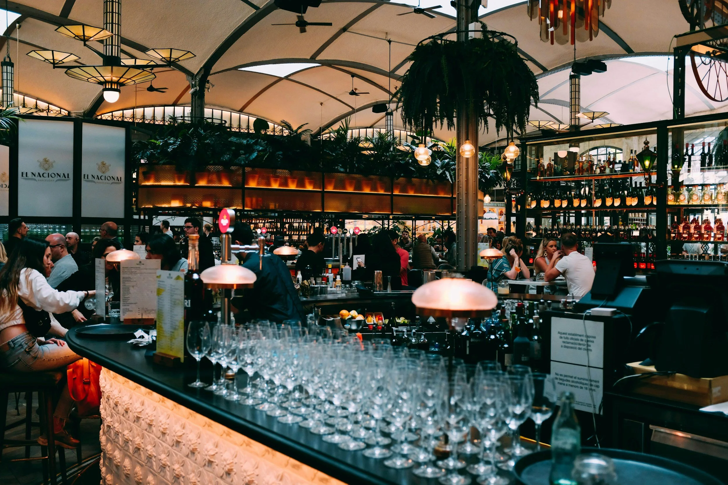 A busy indoor restaurant with many patrons seated at tables and a bar area, decorated with plants, hanging lights, and shelves stocked with bottles.