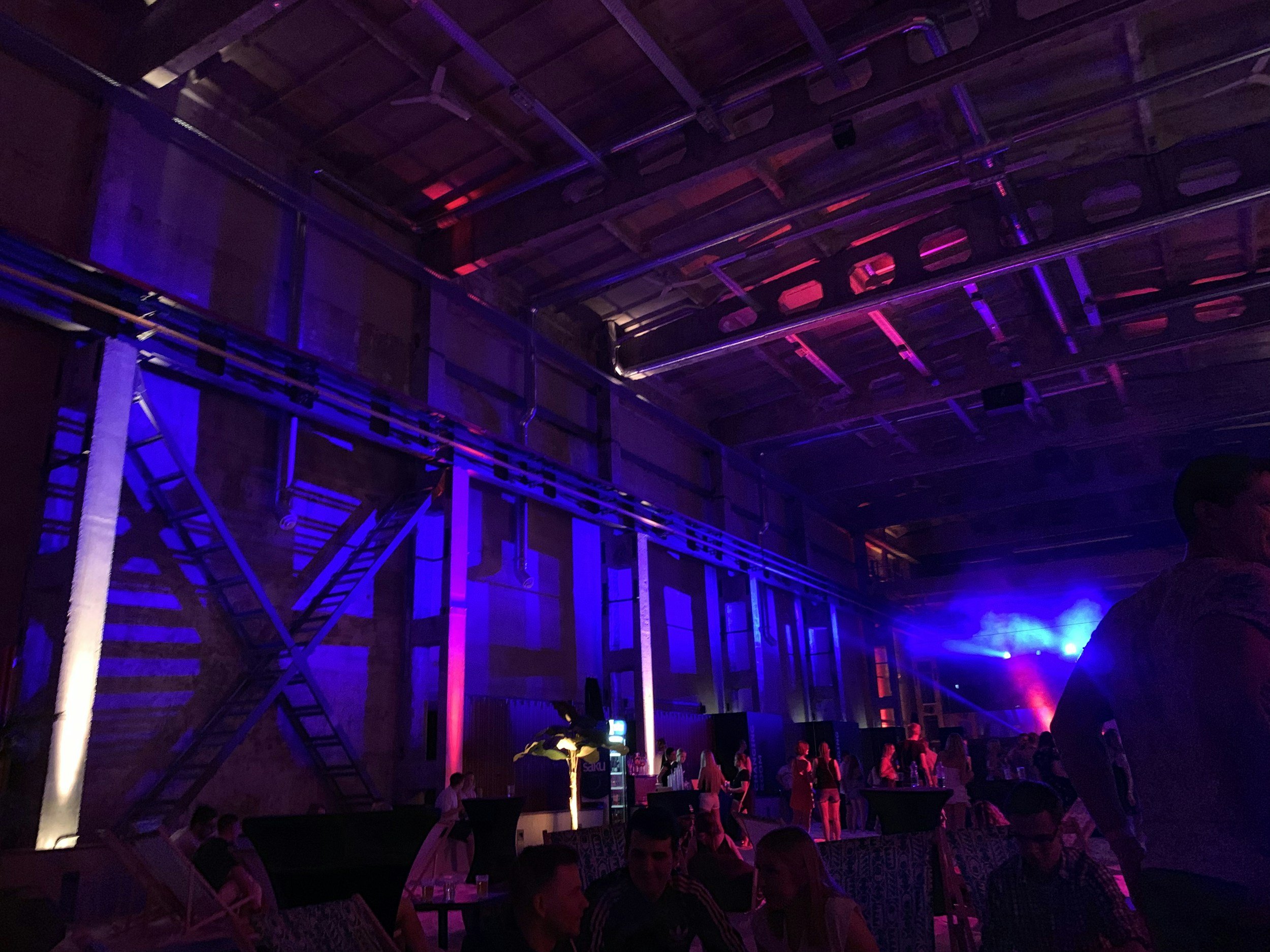 Interior of a nightclub with vibrant purple and pink lighting, people dancing and socializing, and a high ceiling with exposed industrial elements.