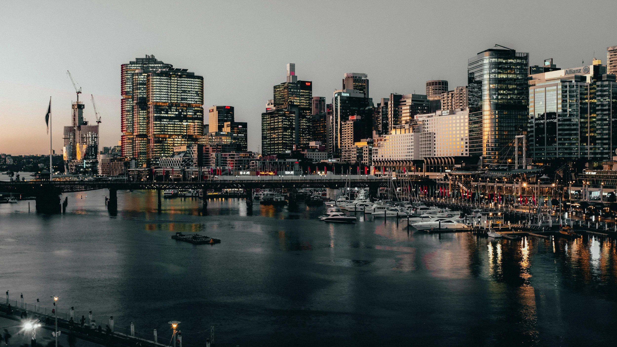 City skyline at dusk with high-rise buildings, marina with boats, and water reflecting city lights.