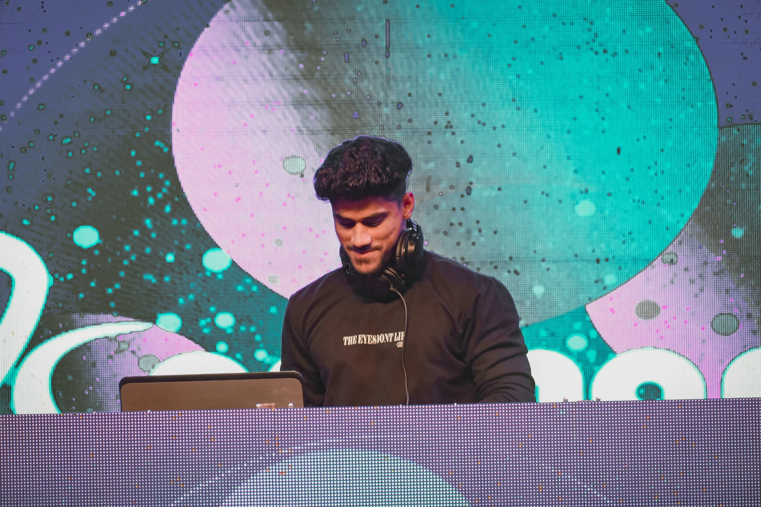 A male DJ with headphones around his neck standing behind a DJ booth, with a colorful digital screen in the background displaying abstract shapes and dots.
