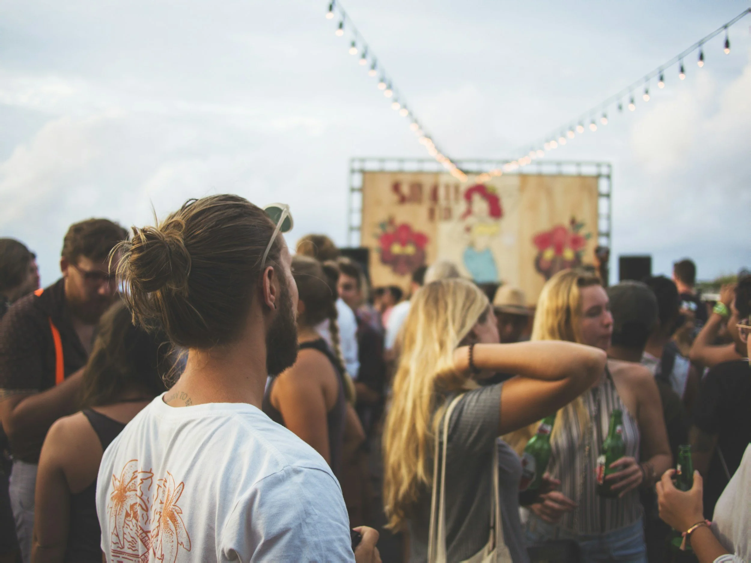 Crowd of people at an outdoor event with string lights and a stage backdrop featuring a woman in a blue dress and red flowers.