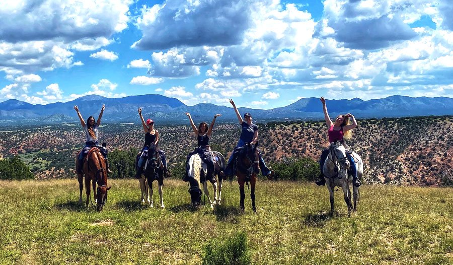 Five people riding horses in a grassy field with mountains in the background under a partly cloudy sky.