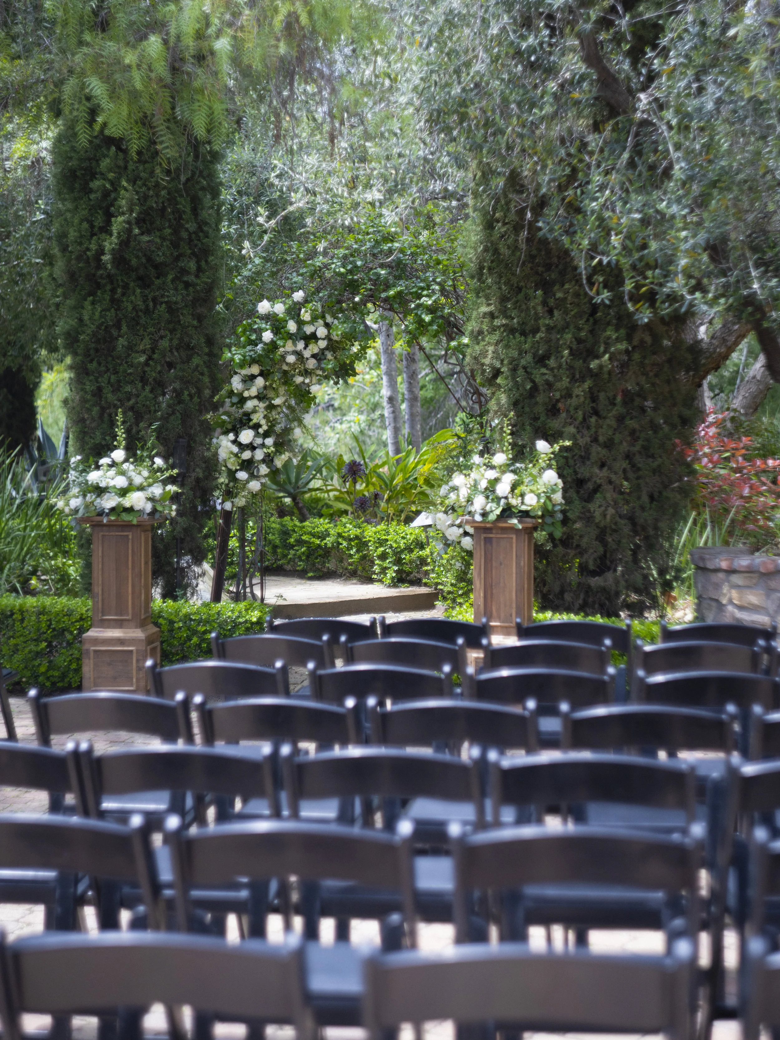 Outdoor wedding ceremony setup in a garden with wooden chairs arranged in rows, white floral arch and pedestals with white flowers, surrounded by green trees and plants.