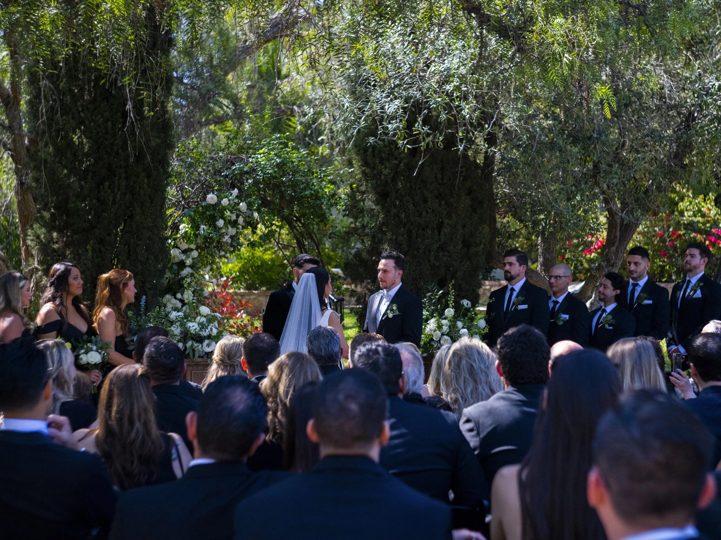 A wedding ceremony outdoors with the bride and groom facing each other, surrounded by bridal party and guests, in a lush garden setting with trees and floral arrangements.