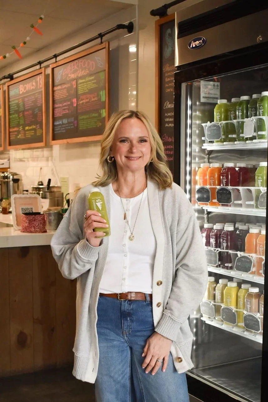 A woman with blonde hair, wearing a cream-colored sweater and ripped jeans, sitting at a table and holding a green juice bottle.