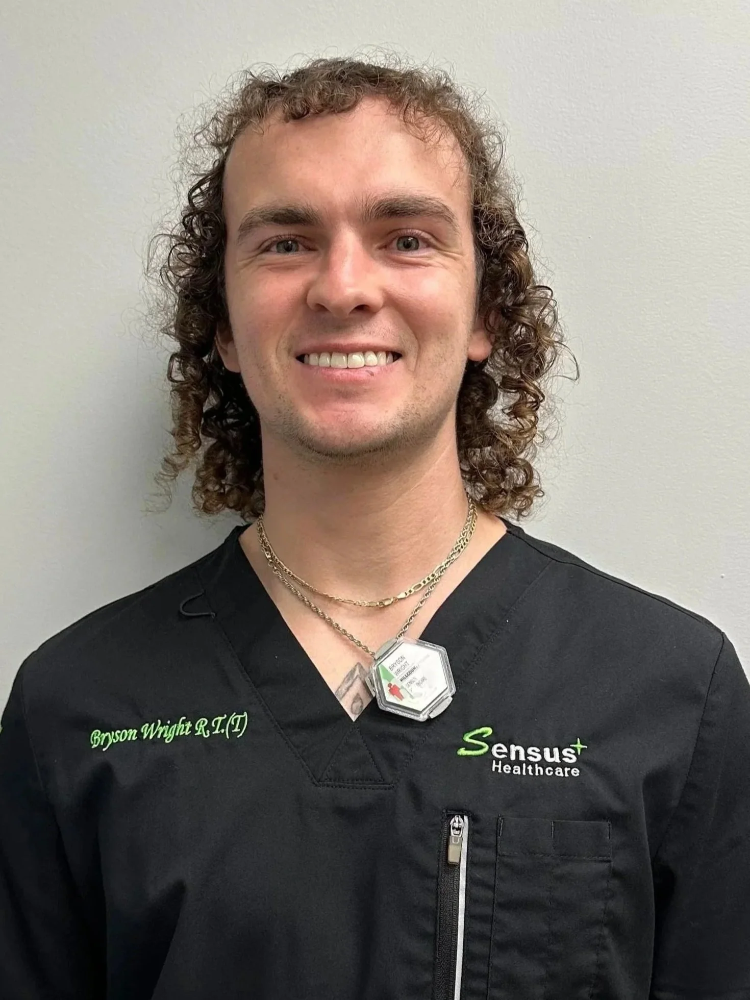 A smiling man with curly brown hair wearing a black medical scrub top with the name 'Bryson Wright R.T(T)' and 'Sensus Healthcare' embroidered on it, standing against a plain off-white wall.